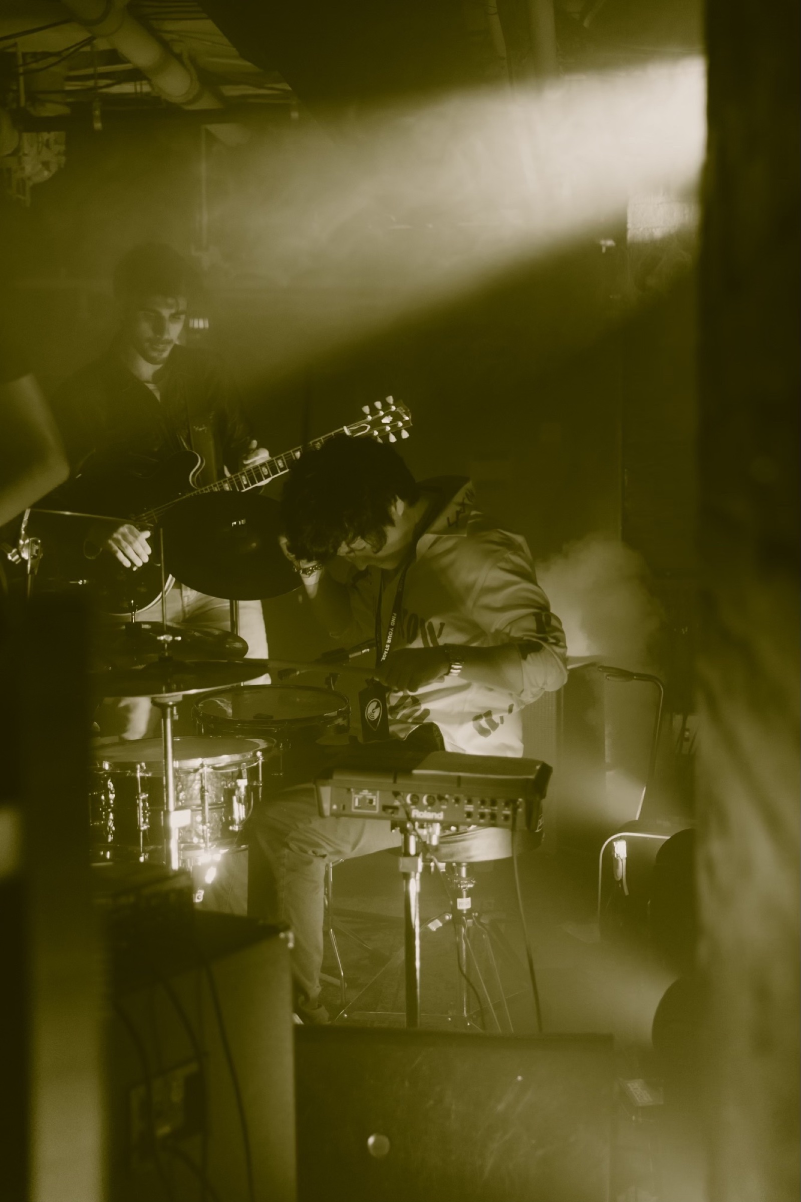 Mir Hwang playing drums in a smoky club, lit warm against a guitarist