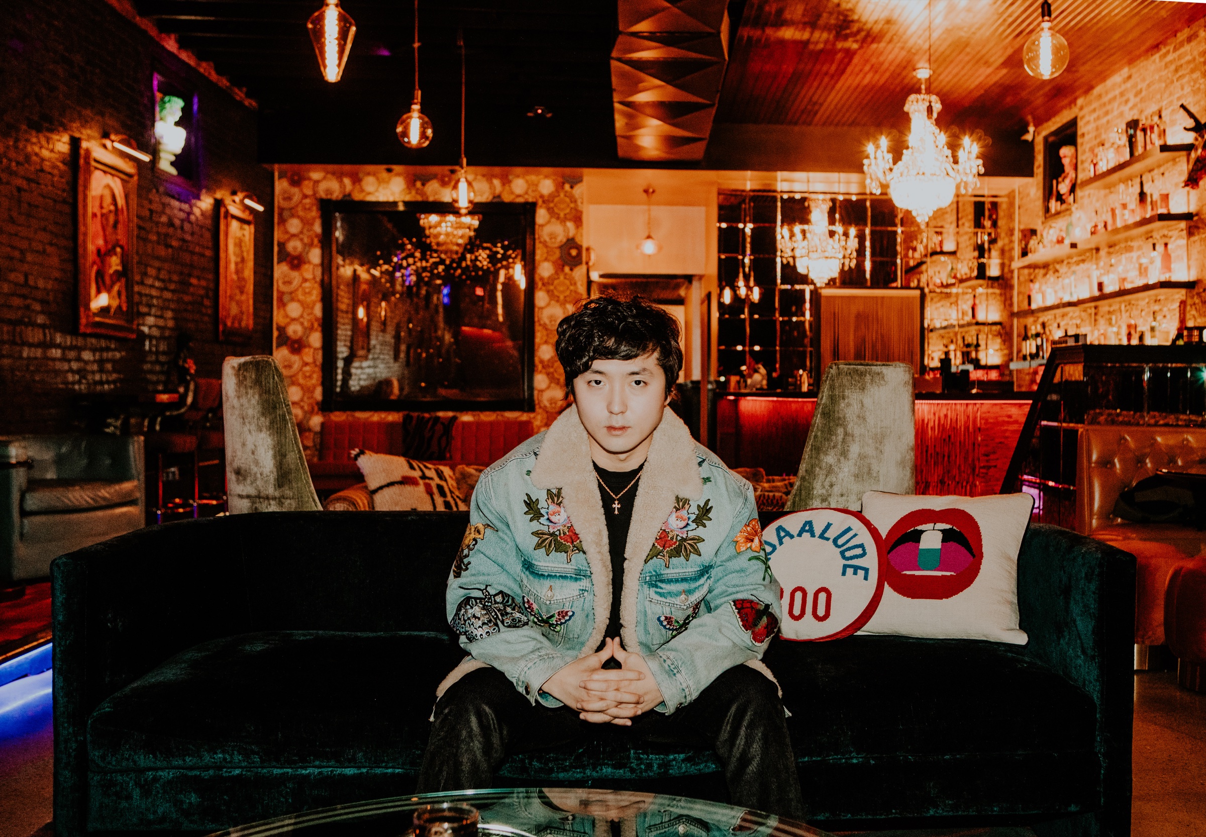 Mir Hwang seated on a velvet couch in a candlelit lounge, wearing an embroidered shearling jacket